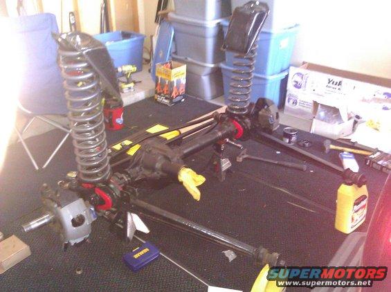 Vehicle axles, springs, and suspension parts laid out on a table. Some parts have yellow rags stuffed in them