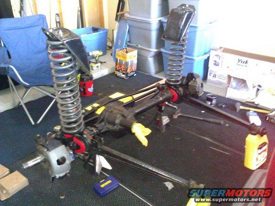 Front axle with coil springs and shock mounts on a dark surface, with tools and storage bins in the background