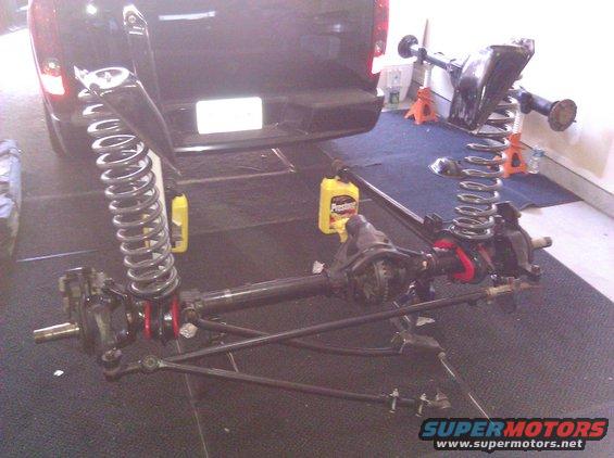 Disassembled front axle, coil springs, and suspension parts laid out on the floor, with the rear of a black SUV visible in the background