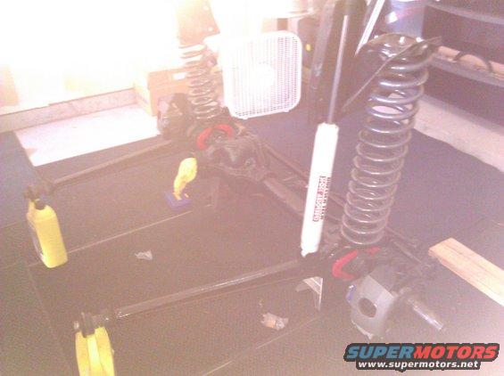 Vehicle suspension parts, including shocks and springs, laid out on a dark surface. A white electric fan is in the background
