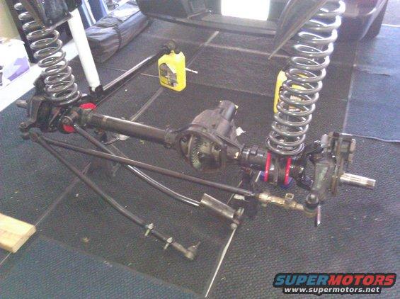 Front axle assembly with coil springs, shocks, and steering linkage. Yellow fluid bottle in background
