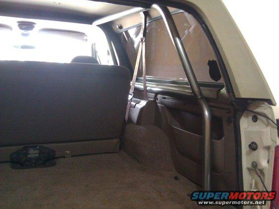 Interior rear of a vehicle showing a roll cage, brown upholstery, and a seatbelt