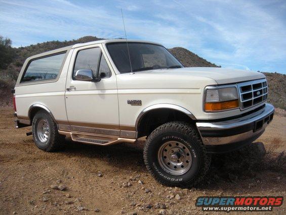 White Ford Bronco parked on dirt road with hills in background. Has chrome accents and running boards