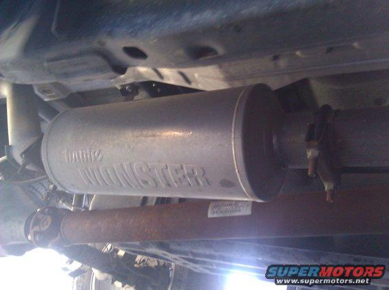Underside of a vehicle showing a silver exhaust muffler with 'MONSTER' embossed on it, and a rusty driveshaft