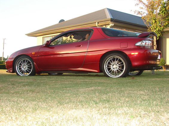 Side view of a red coupe with aftermarket wheels parked on a lawn in front of a house