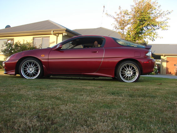 Red 1993 Mazda MX-3 coupe parked on grass in front of a house