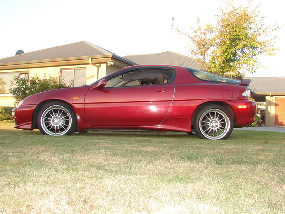 Side view of a red coupe with aftermarket wheels parked on a lawn in front of a house