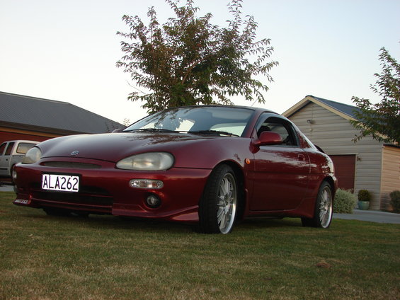 Red coupe parked on grass in front of a garage. License plate ALA262