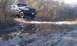 1992 Ford Bronco - A 1992 Ford Bronco navigates a challenging muddy terrain with a significant puddle reflecting the sky and surrounding brush.