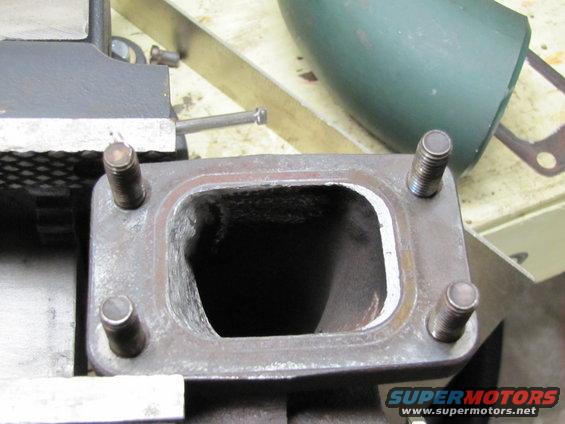 Close-up of a turbo exhaust manifold flange with four studs. A green pipe is in the background