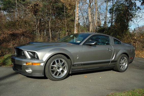 Gray Ford Mustang GT500 parked on asphalt with trees in background. Side view shows GT500 badging