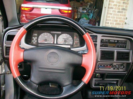 Isuzu interior with red and black steering wheel, dashboard gauges, and radio. Red SUV visible outside