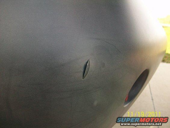 Close-up of a black plastic bumper with a slit and a circular cutout, showing a date and website watermark