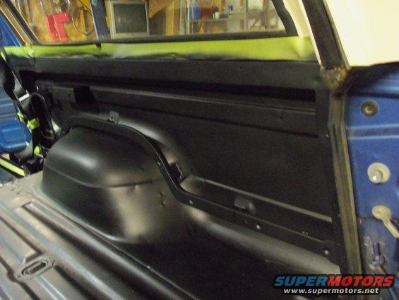 Interior of a blue vehicle, showing the rear wheel well and floor pan, with green masking tape on the upper trim