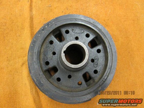 Close-up of a black crankshaft pulley with holes and a metal center. Text and a date stamp are visible