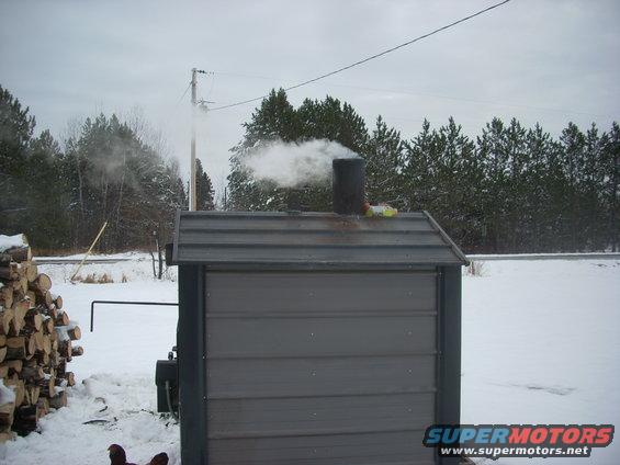 Wood boiler with smoke from chimney, pile of logs, snowy ground, trees, utility pole
