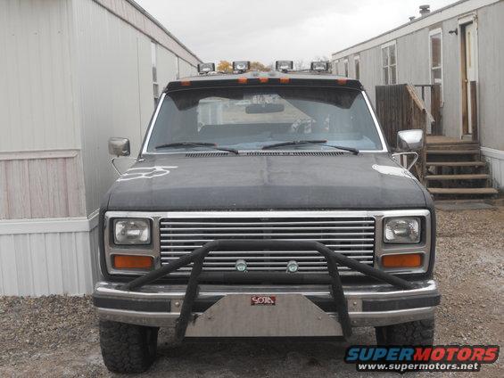 Front view of a dark-colored Ford Bronco with a brush guard and roof lights. It's parked in front of some trailers