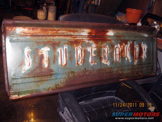 Rusty tailgate with "STUDEBAKER" embossed in letters, showing teal and red paint