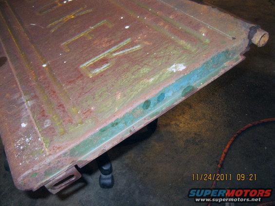 Close-up of a rusty metal panel with embossed letters that appear to spell 'TRUCK'. It has a green and blue painted edge