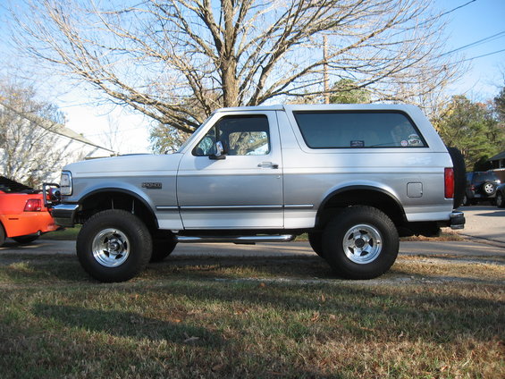 1994 Ford Bronco parked on grass, side view. Orange car visible to the left, other vehicles in background
