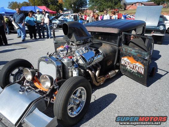 Rat rod with a large chrome engine, black roof, and "Mountain Magic Speed Shop" painted on the driver's door