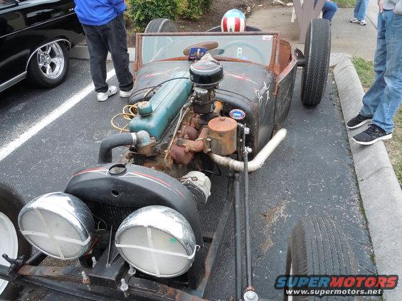 Rat rod with a big engine, exposed front end, and a helmet on the dash. Another car is parked next to it