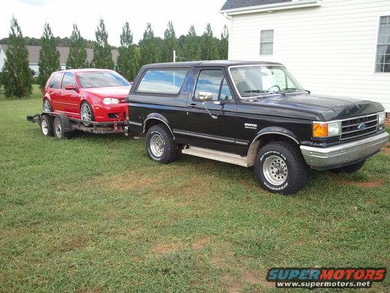 Black Ford Bronco towing a red car on a trailer, viewed from the front 3/4 angle