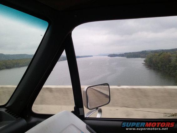 Interior of a vehicle with a side mirror reflecting a road, looking out at a wide river and tree-lined shores under a cloudy sky