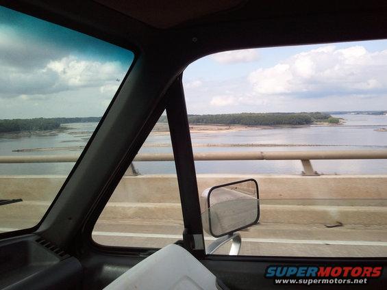 Interior view from a vehicle, looking out the passenger side window at a river and bridge. Watermark reads SUPERMOTORS