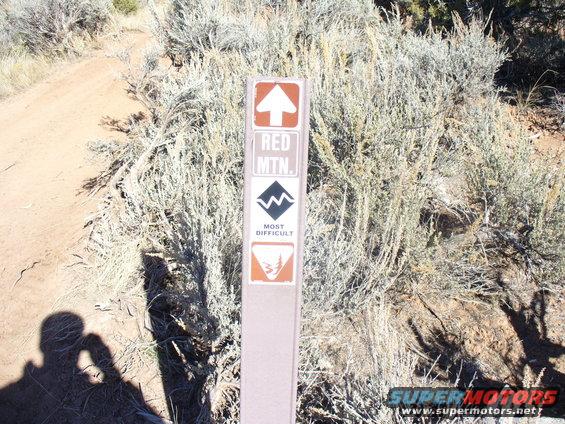 Trail marker sign with 'RED MTN.' and 'MOST DIFFICULT' text, surrounded by dry brush