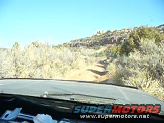 Driving on a dirt road through a dry, brushy landscape under a clear blue sky. The dashboard and windshield are visible in the foreground
