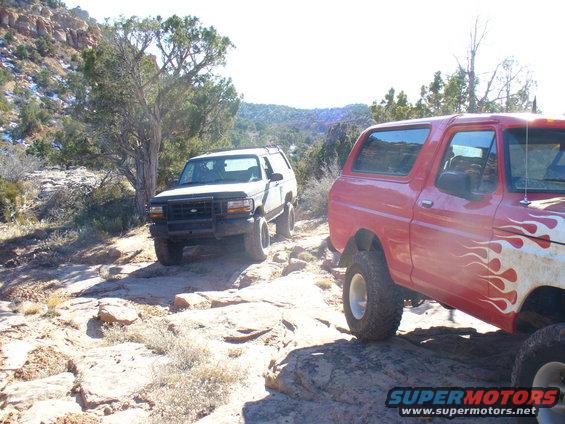 White SUV from the front-left, red SUV from the rear-right on a rocky, arid trail