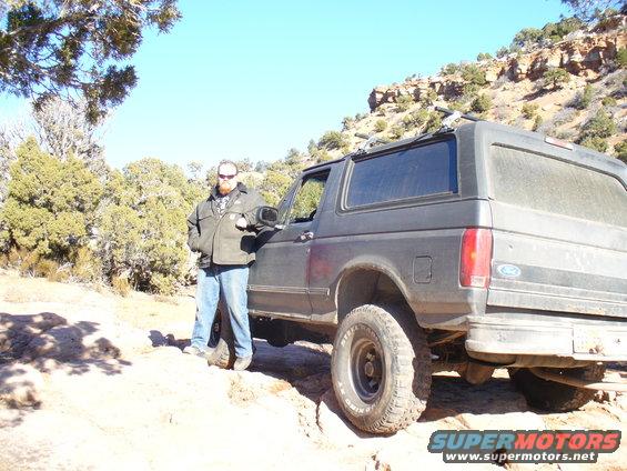 Guy leaning on a dark gray Ford Bronco parked on a dirt trail with rocky hills and trees in the background