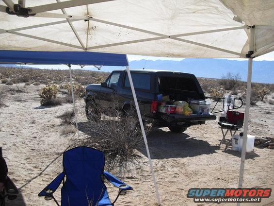 Black Ford Bronco with tailgate open, parked in a desert campsite under pop-up canopies. Camping gear is visible in the cargo area
