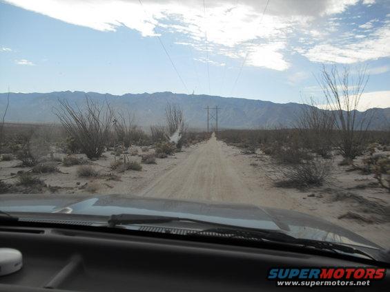Dashboard and windshield view from inside a vehicle driving on a dusty dirt road through a desert with mountains in the distance