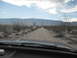 1986 Ford Bronco - View from inside a vehicle on a dirt road in a desert with mountains and power lines under a cloudy sky.