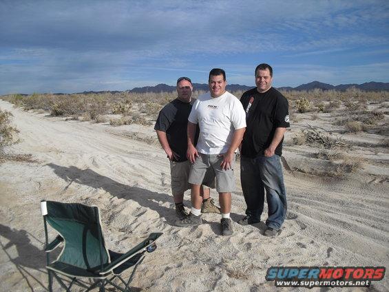 Three men stand in a desert with scrub brush and mountains in the background. A folding chair is in the foreground