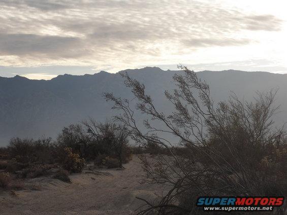Desert landscape with mountains, dry brush, and a dirt path under a cloudy sky. "SUPERMOTORS.NET" text is visible