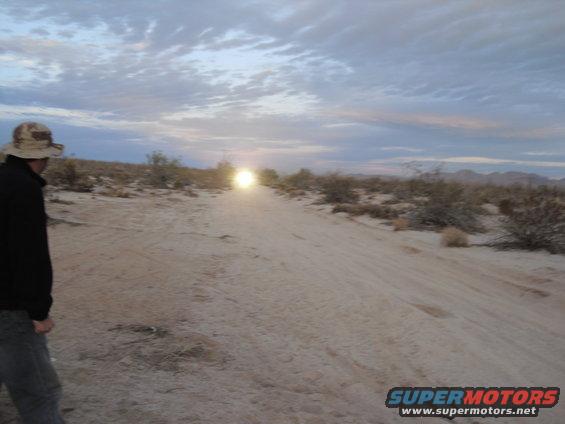 Person in hat looks at distant headlights on a dirt road in the desert at dusk