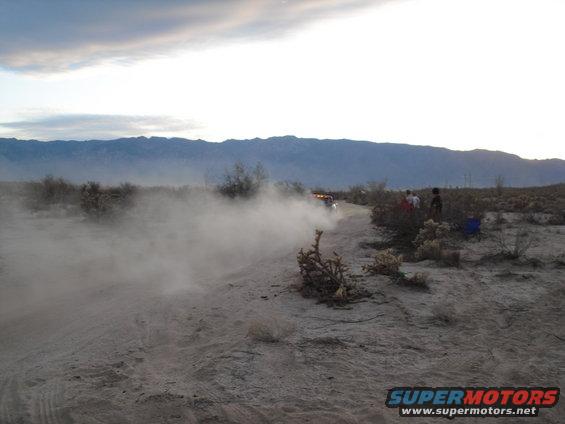 Off-road vehicle driving on a dirt track, kicking up a large dust cloud. Mountains in the background
