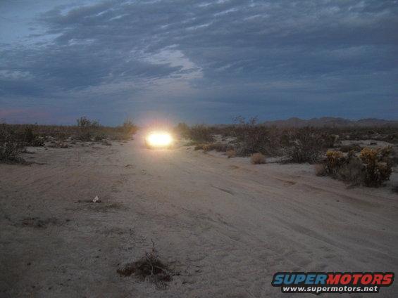 Vehicle driving on dirt road in desert at dusk, headlights on