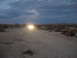 1986 Ford Bronco - A vehicle drives down a dusty desert road as the sun sets, its headlights cutting through the twilight.