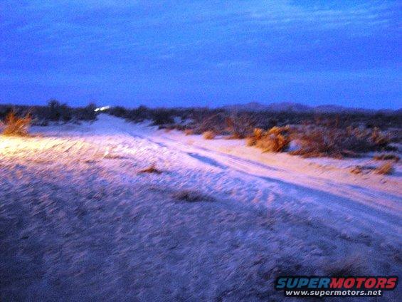 Desert landscape at dusk with tire tracks and distant headlights illuminating the sandy terrain
