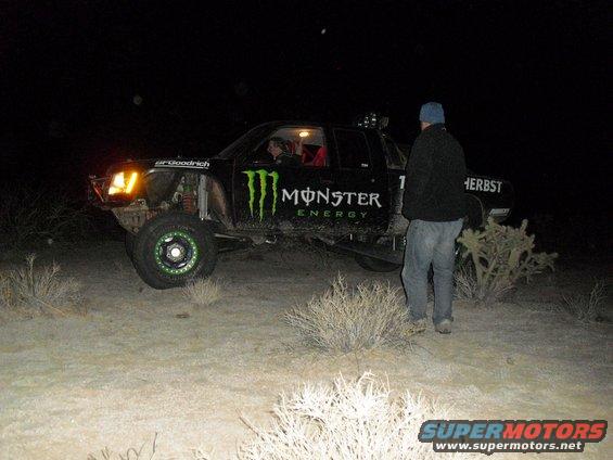 Monster Energy truck with off-road tires and suspension, parked at night in a desert area. A person stands nearby