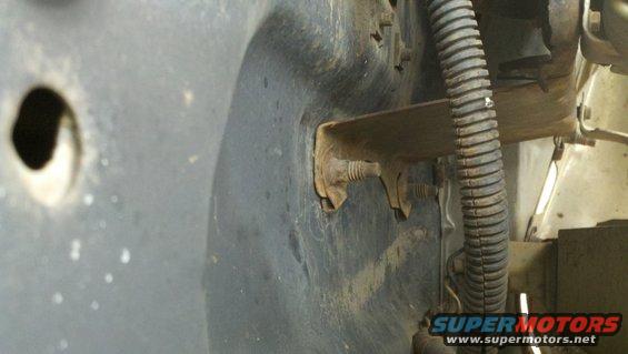 Close-up of a rusty metal bracket bolted to a dark, possibly plastic, fender liner with a hole in it. Corrugated wire loom is visible