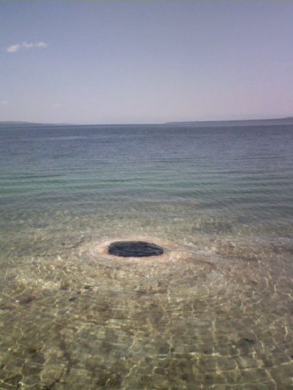 Circular dark hole in shallow, clear water with ripples. Distant landmass on horizon under a pale blue sky