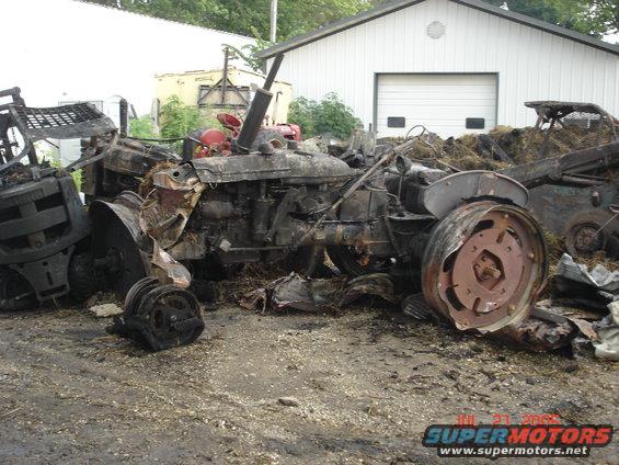 Burnt tractor with red engine components visible, parked in front of a white garage