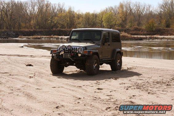 Jeep Wrangler parked on a sandy area near a body of water with trees in the background