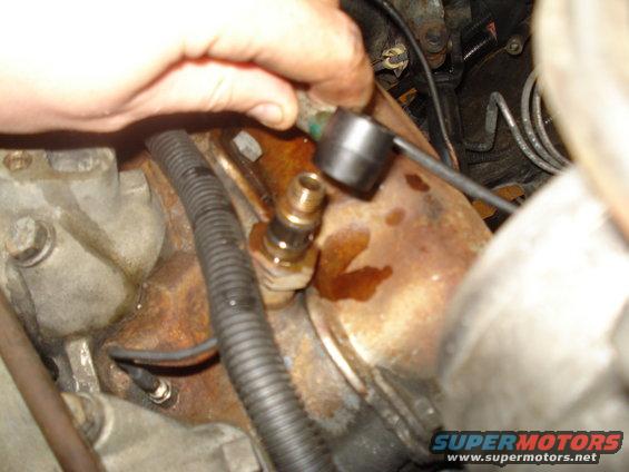 Hand holding a tool on a brass fitting screwed into a rusty engine component, with black wiring loom nearby
