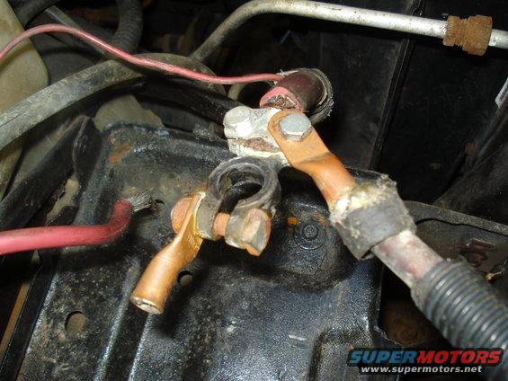 Corroded battery terminals with copper cables connected to a black mounting bracket. Red wire visible